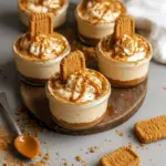 Close-up of No-bake Biscoff Cheesecake Cups in clear glasses showing crumbly Biscoff crust, creamy cheesecake filling, and a shiny cookie-butter swirl topped with a halved biscuit.
