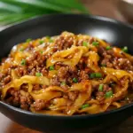 Skillet of glossy Potsticker Noodle Bowl with ground pork, wide noodles, and bright cabbage slaw, garnished with green onions—an inviting example of quick Potsticker Noodles.