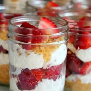 Glass jar of Healthy No-Bake Strawberry Cheesecake showing vanilla cookie crumbs, a pale pink yogurt-cheesecake layer, and fresh strawberry slices on top.
