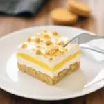 Slice of Lemon Oreo Dessert on a white plate: golden-Oreo crumb crust, bright yellow lemon pudding, and fluffy whipped topping dusted with cookie crumbs.