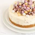 Slice of No-Bake Mini Egg Cheesecake on a white plate: crunchy biscuit base, creamy cheesecake center, and colorful Mini Eggs piled on top.