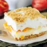 Close-up of No-Bake Peach Yum Yum in a glass dish: golden graham crust, fluffy cream-cheese layers, glossy peach pie filling, and a light sprinkle of crumbs.