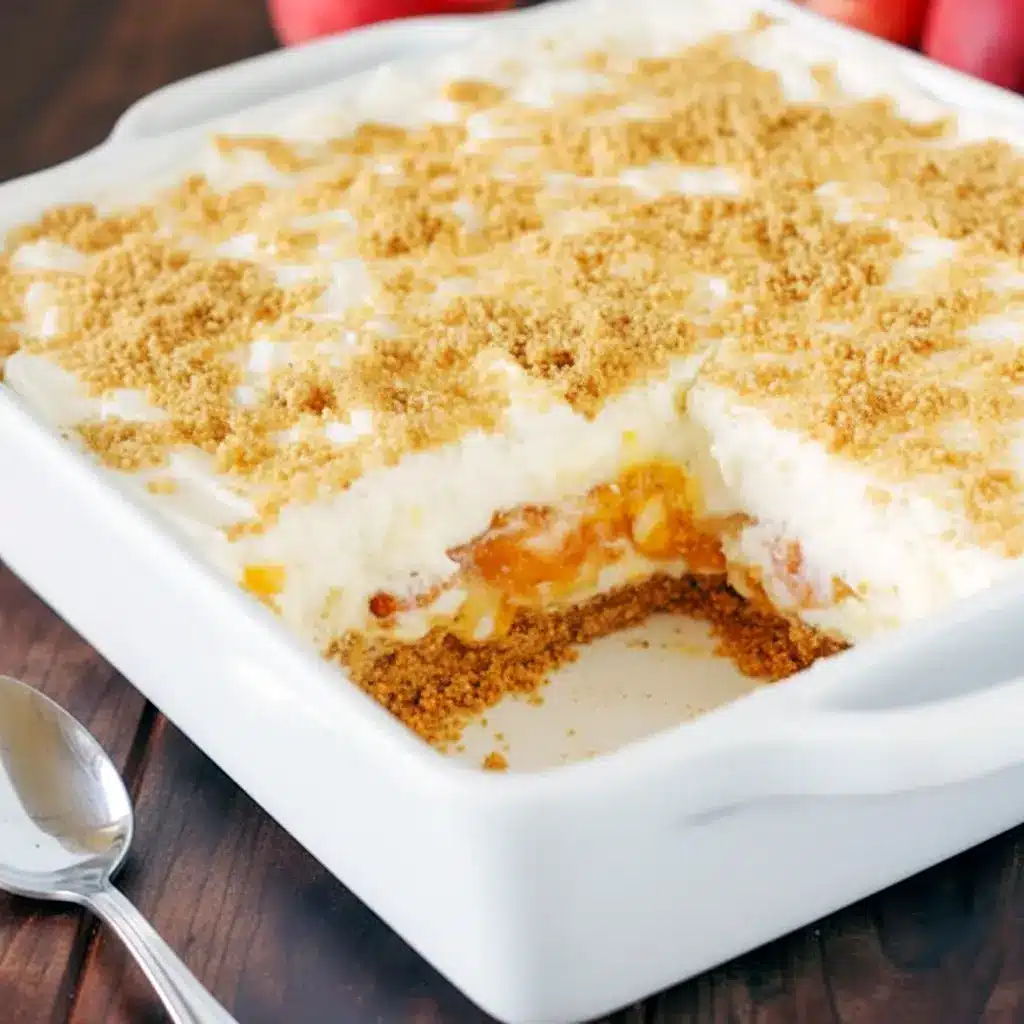 Close-up of No-Bake Peach Yum Yum in a glass dish: golden graham crust, fluffy cream-cheese layers, glossy peach pie filling, and a light sprinkle of crumbs.