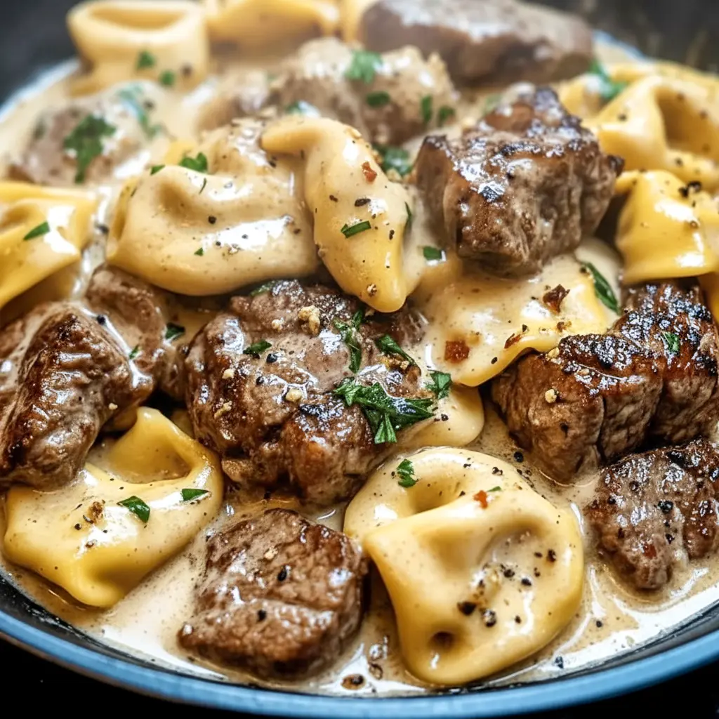 Garlic Steak bites and cheesy Tortellini in a creamy sauce in a hot Skillet, garnished with parsley.