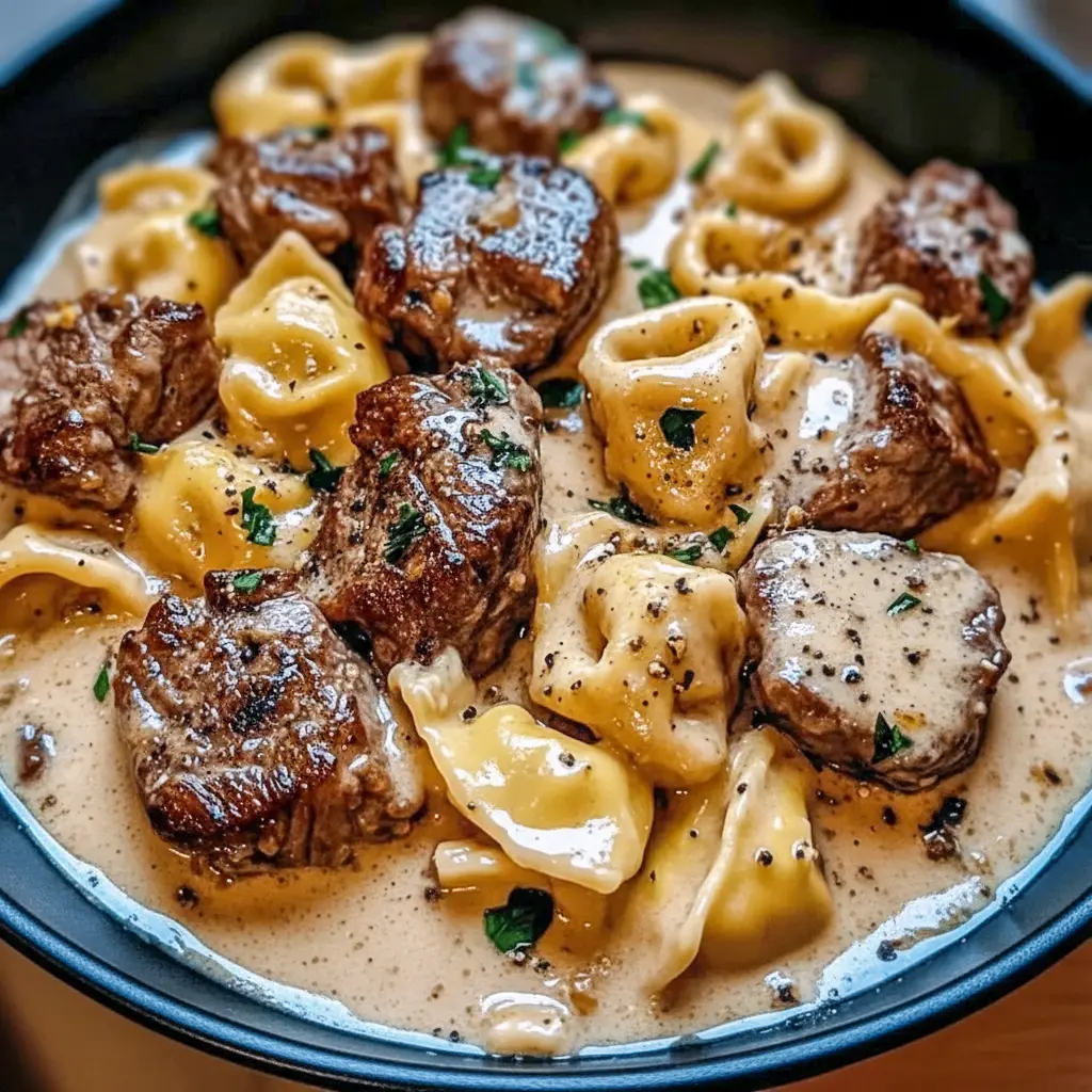Garlic Steak bites and cheesy Tortellini in a creamy sauce in a hot Skillet, garnished with parsley.